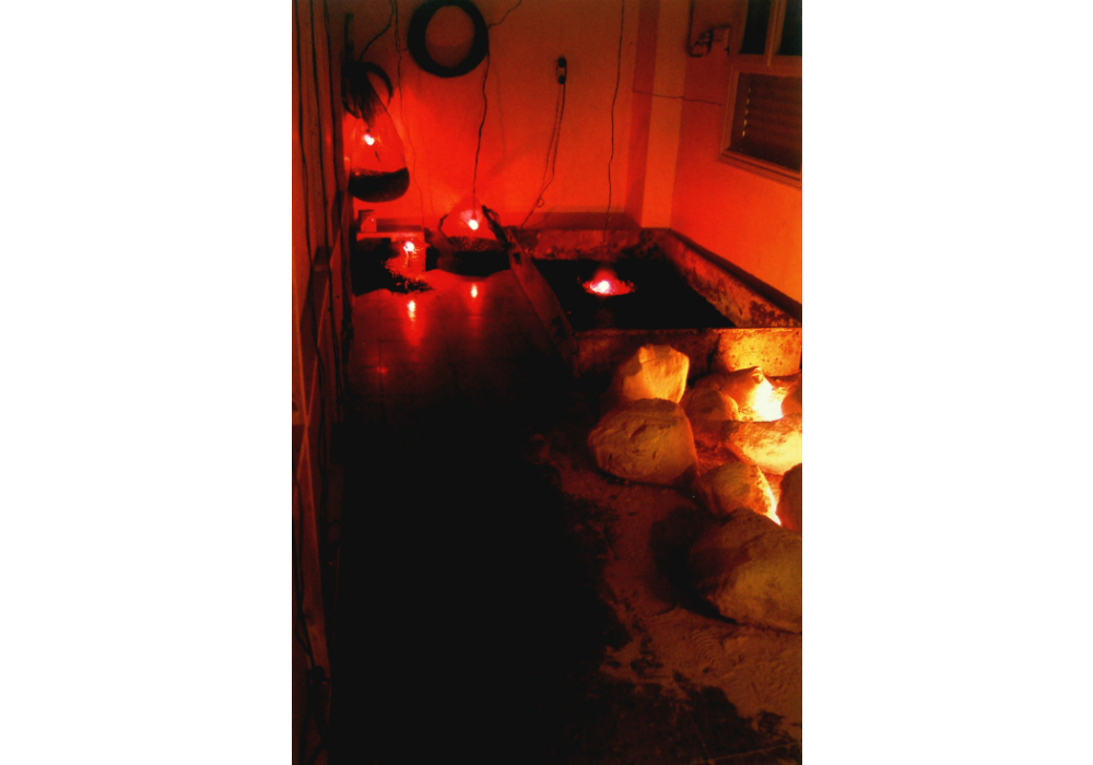Behind the Wall (2004),
installation, discarded construction-site materials, black olive pits and dried sand bags, barrel lids with photographs, Jerusalem scrolls red and yellow light
Behind the wall